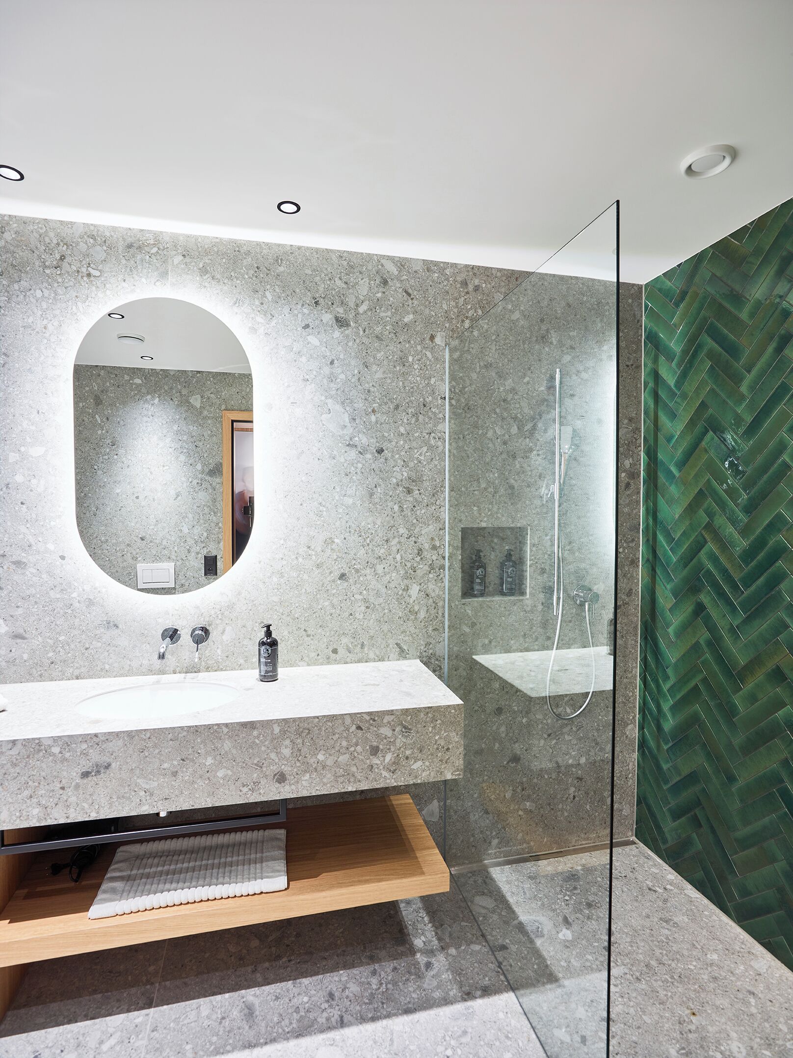 A modern bathroom with a granite countertop, a wooden shelf, a glass shower enclosure, and a herringbone-patterned wall.