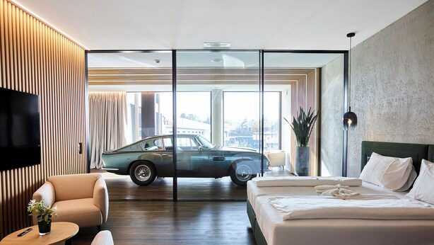 Una habitación de hotel moderna con una cama grande, un sofá, una mesita y un televisor. Un coche clásico está estacionado fuera de la ventana.