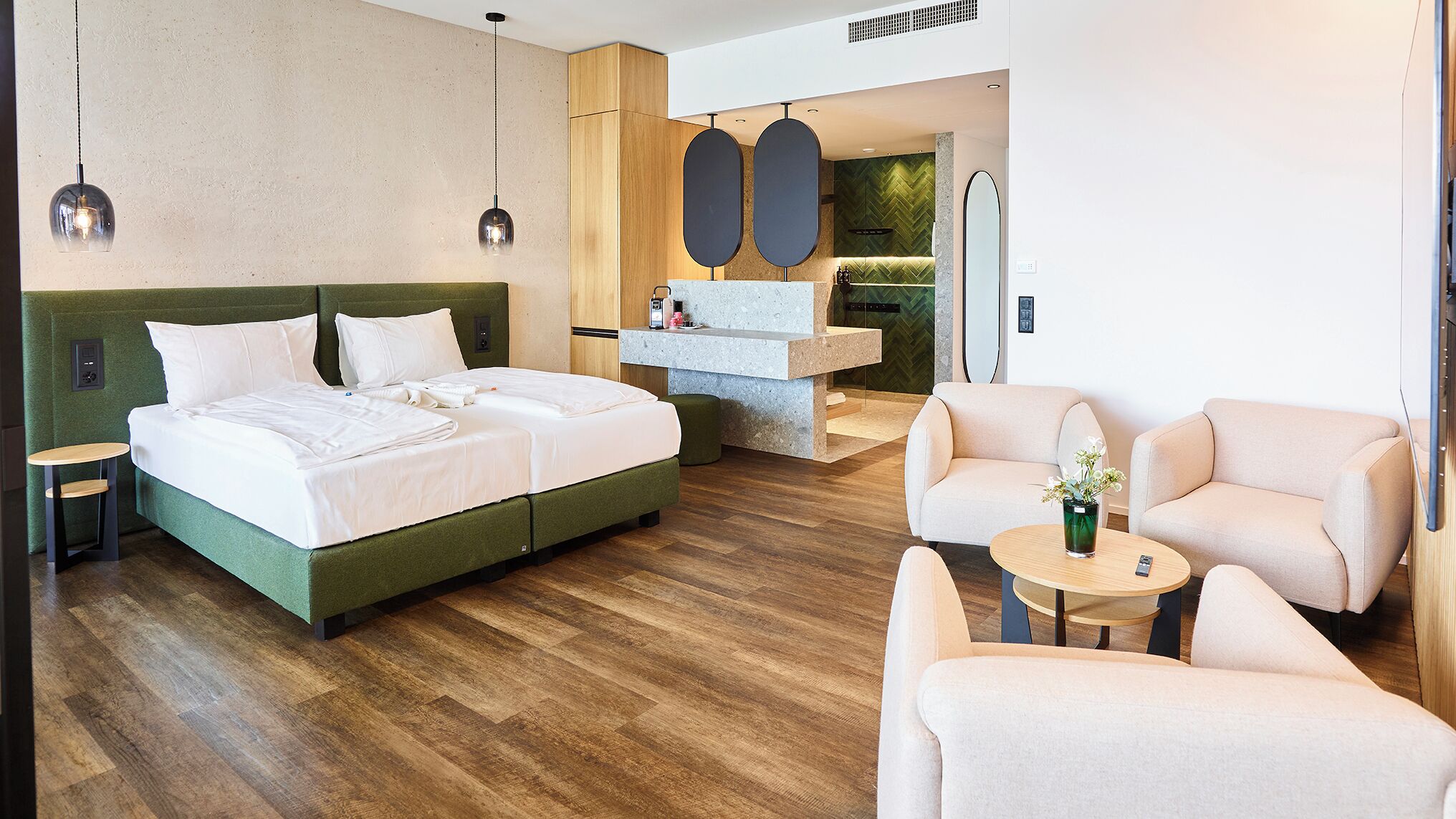 Ein modernes Hotelzimmer mit einem großen Bett, einer Sitzecke mit zwei Stühlen, einem kleinen Tisch und einem im Hintergrund sichtbaren Badezimmer.