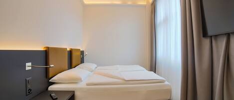 A neatly made bed with white linens, a bedside table with a remote control, and a wall-mounted light fixture.