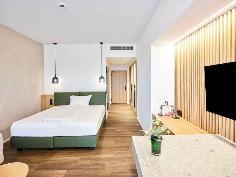 A modern hotel room with a bed, bedside table, TV, and a small kitchenette area.