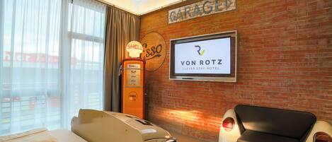 Habitación de hotel con pared de ladrillo, una gasolinera Shell, un televisor mostrando el nombre del hotel y un coche antiguo.