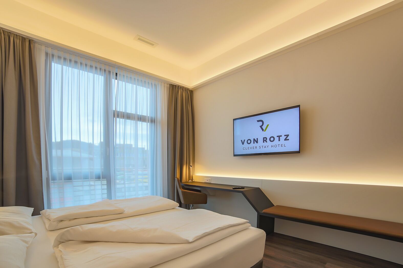 Ett hotellrum med en säng, ett skrivbord och en tv som är monterad på väggen och visar hotellets namn.