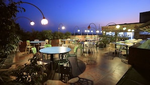 Un patio al aire libre con mesas y sillas redondas, rodeado de plantas en macetas.