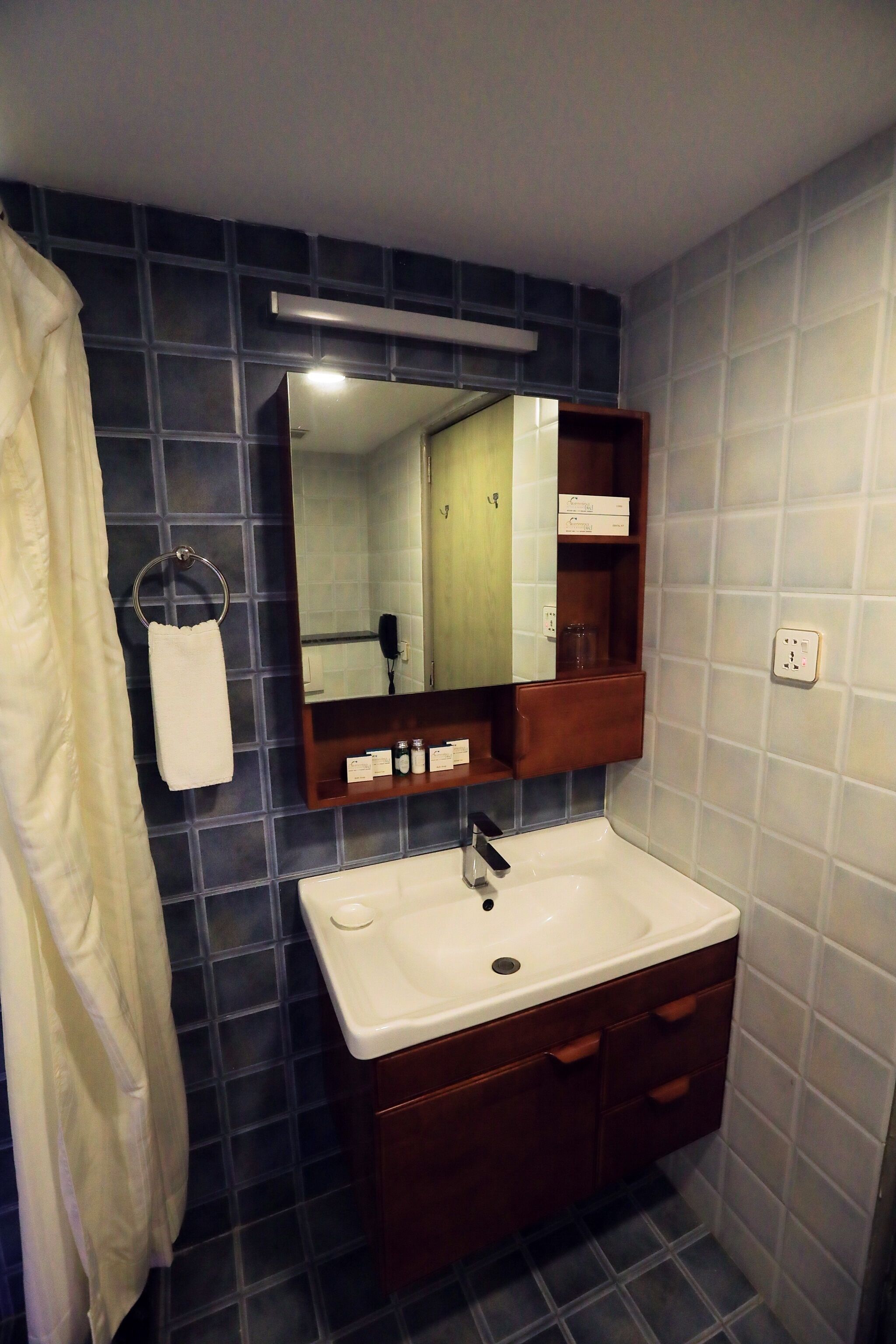 A bathroom with a white sink, dark blue tiled walls, a wooden vanity, a mirror, and a towel hanging on a rack.