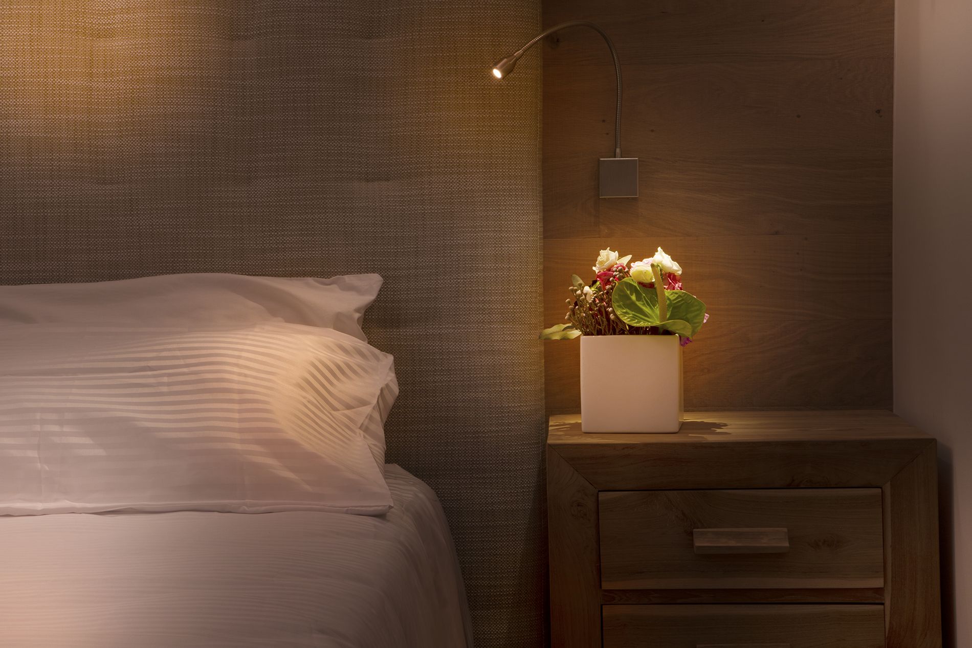 A neatly made bed with white linens, a bedside table with a potted plant, and a wall-mounted lamp.