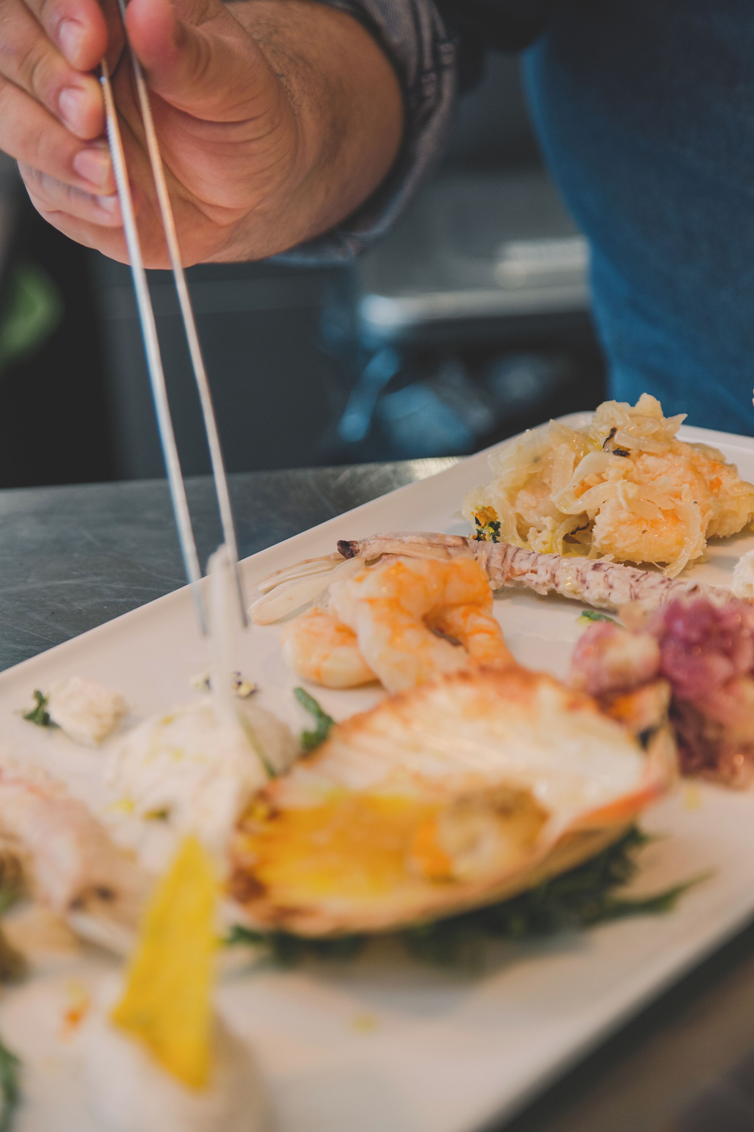 A person holding a pair of tongs over a plate of food, including shrimp and a piece of grilled fish.