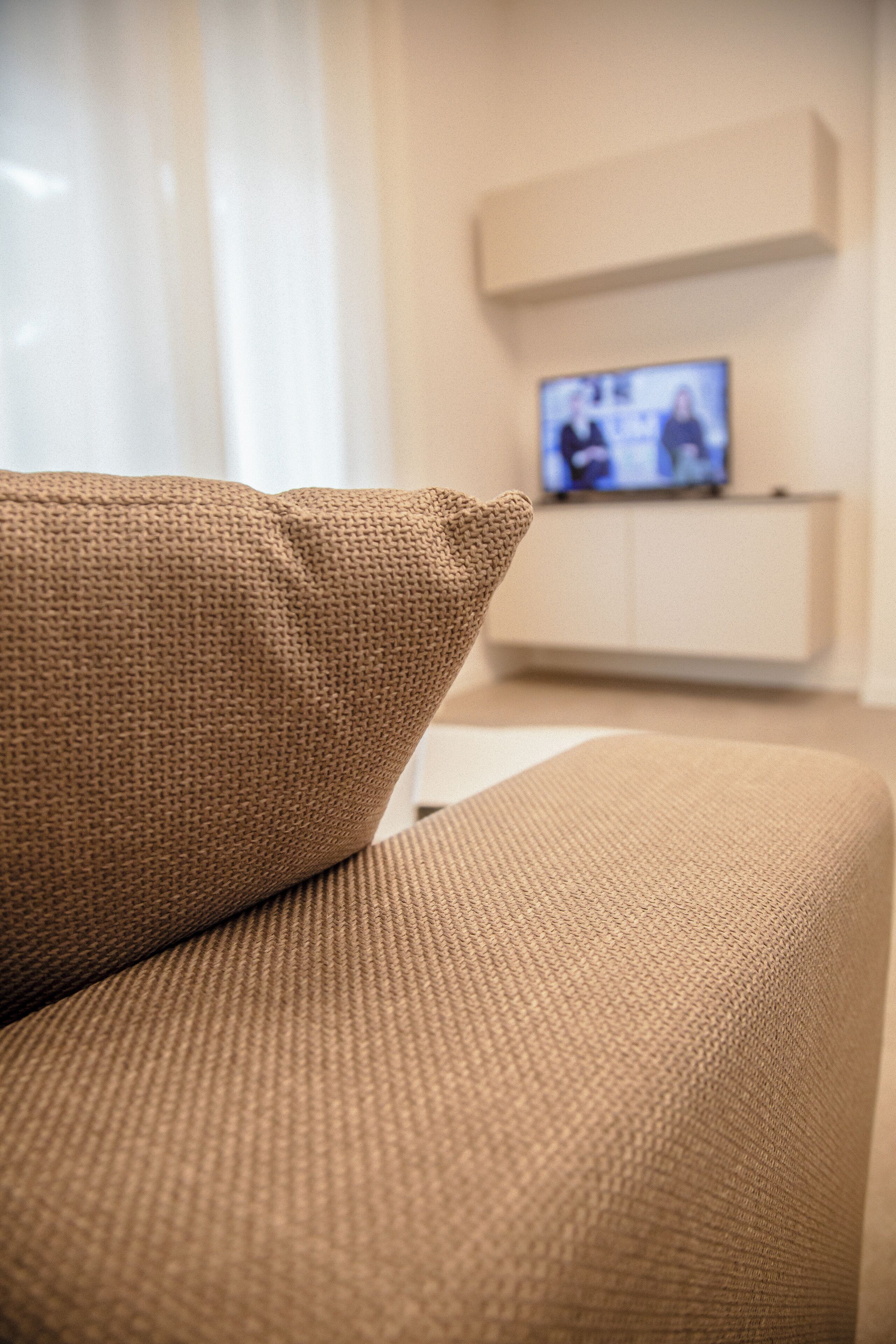 Un sofá color beige con tela texturizada, un televisor sobre un mueble en el fondo y una pared blanca.