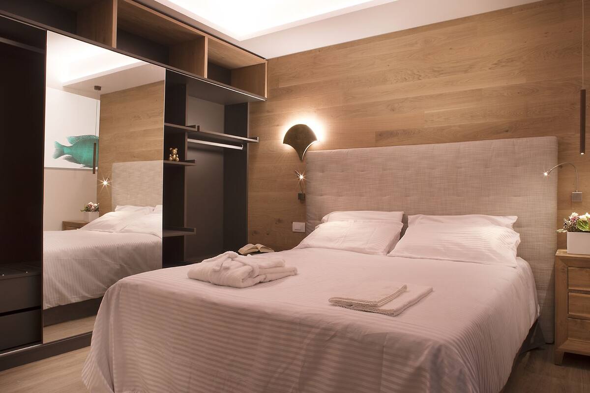 A modern bedroom with a large bed, a wardrobe, and a bedside table with a lamp.