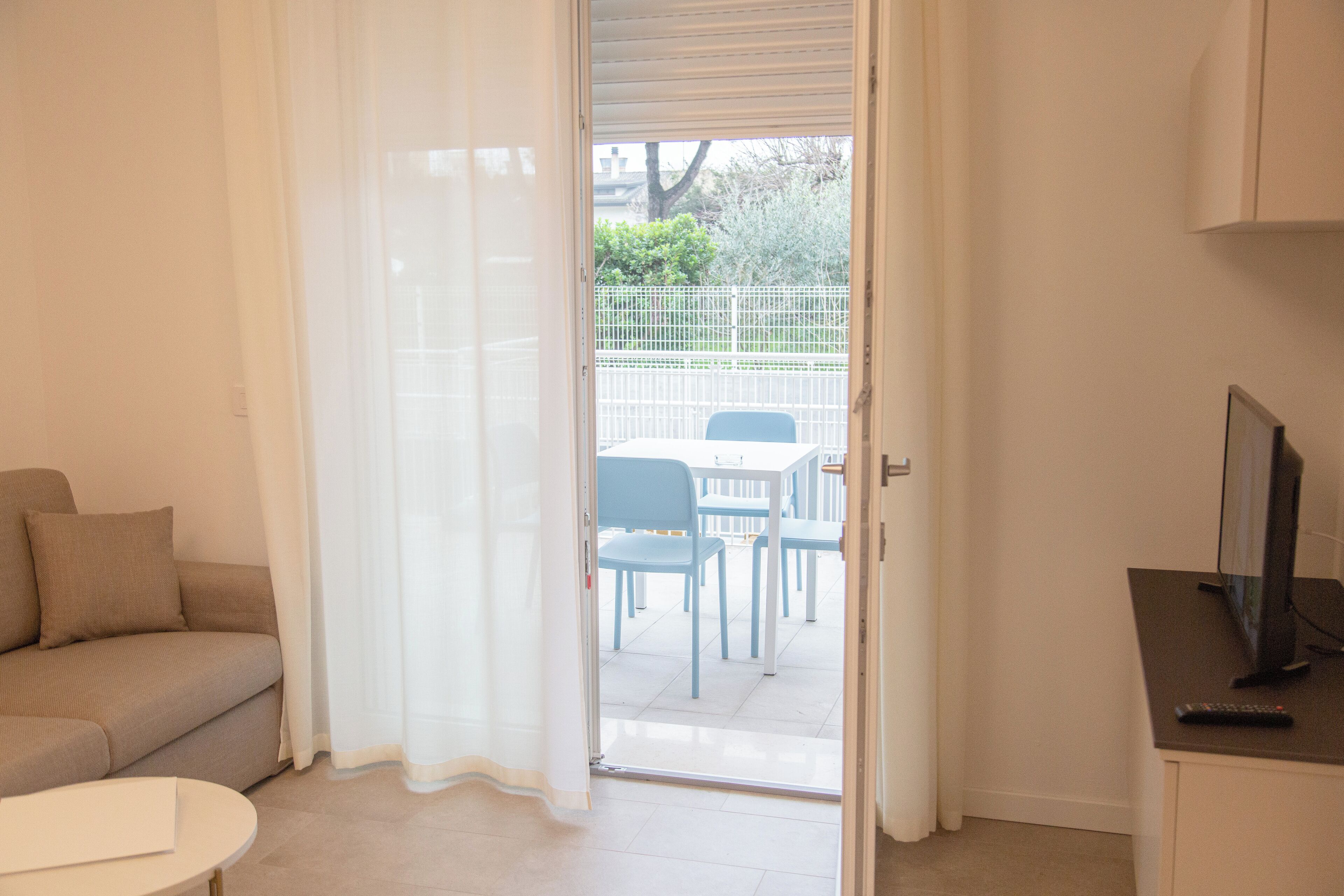 Una habitación con un sofá color beige, una mesita redonda pequeña, un televisor y una puerta que da a un patio exterior con valla blanca y sillas azules.