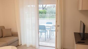 Una habitación con un sofá color beige, una mesita redonda pequeña, un televisor y una puerta que da a un patio exterior con valla blanca y sillas azules.