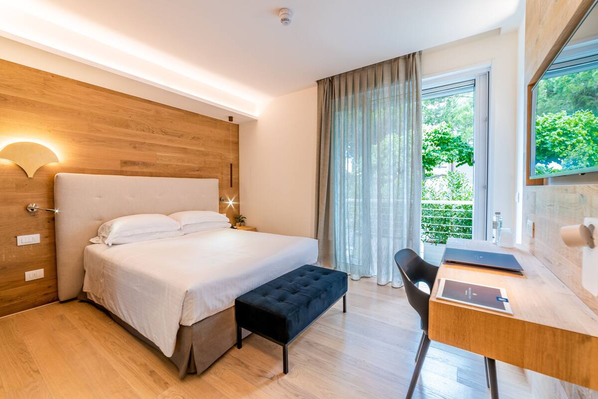 A modern hotel room with a wooden headboard, a large bed, a desk with a chair, and a view of greenery through the window.