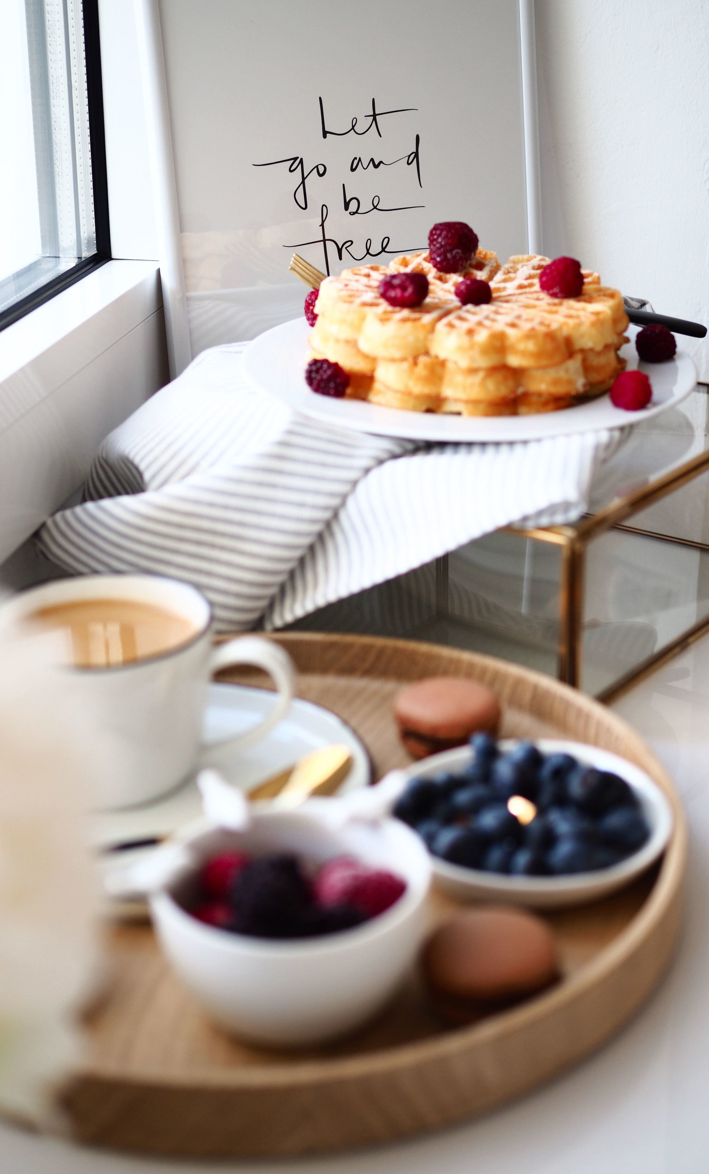 Desayuno con waffles cubiertos de frambuesas, una taza de café y tazones de bayas sobre una bandeja.