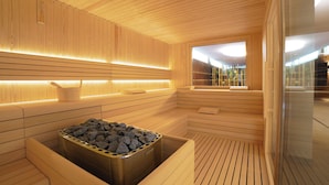 A modern sauna with wooden paneling, a heated stone bath, and a view of the outdoors. - The Green Park Hotel (Cavallino-Treporti)