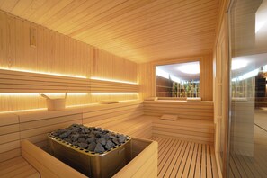 A modern sauna with wooden paneling, a heated stone bath, and a view of the outdoors. - The Green Park Hotel (Cavallino-Treporti)