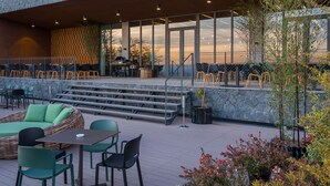 En moderne bygning med en stenfassade, udendørs siddeområde med grønne stole og en trappe, der fører op til en etage.