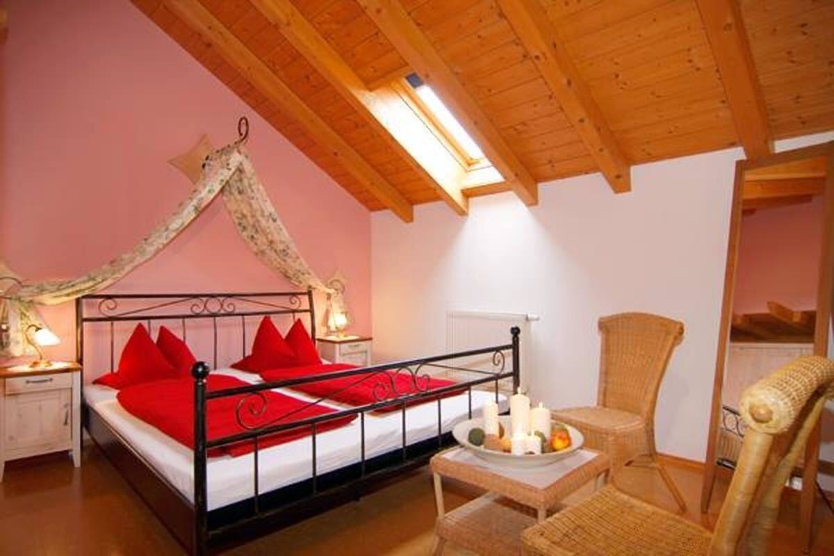 A bedroom with a wooden ceiling, a bed with red bedding, a nightstand, a wicker chair, and a small table with candles.