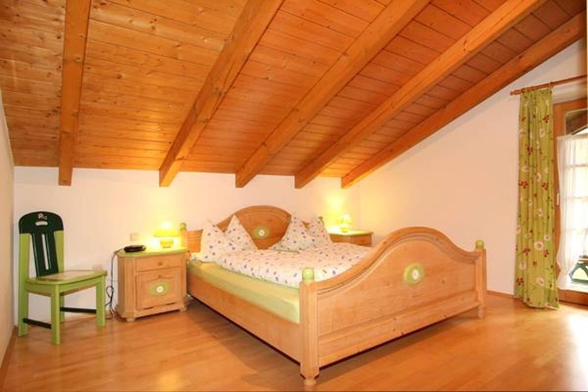 A bedroom with a wooden ceiling, a single bed, a nightstand, a chair, and a window with curtains.