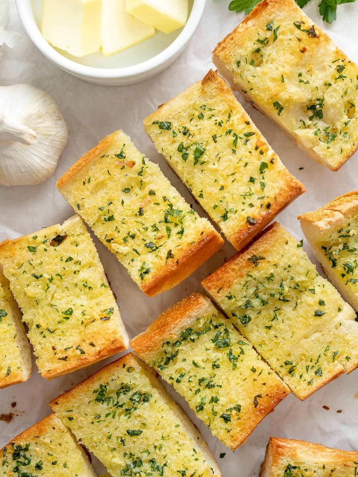 Rebanadas de pan con ajo, adornadas con hierbas, un bol de mantequilla y un ajo entero.