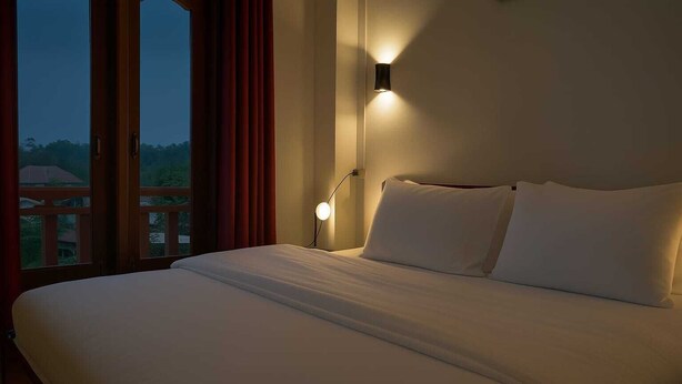 A hotel room with a large bed, a nightstand, a wall-mounted lamp, and a window with curtains. - Sipanya Guesthouse (Phonsavan)