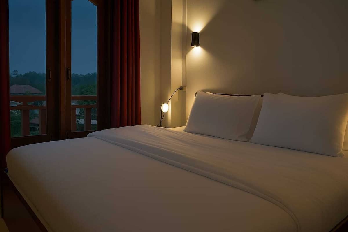 A hotel room with a large bed, a nightstand, a wall-mounted lamp, and a window with curtains.