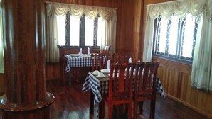 A dining area with wooden paneled walls, large windows with curtains, and tables set with chairs. - Sipanya Guesthouse (Phonsavan)