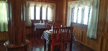 Un comedor con paredes de paneles de madera, ventanas amplias con cortinas y mesas dispuestas con sillas.