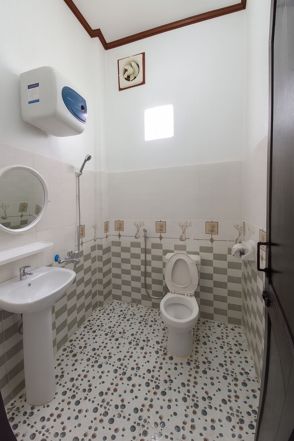 Baño con inodoro, lavamanos y espejo. Cuenta con cabezal de ducha y secador de manos. El piso tiene un diseño de baldosas con motivos.