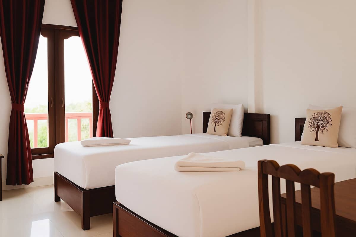 A hotel room with two beds, a wooden chair, a table, and a view of greenery through the window.