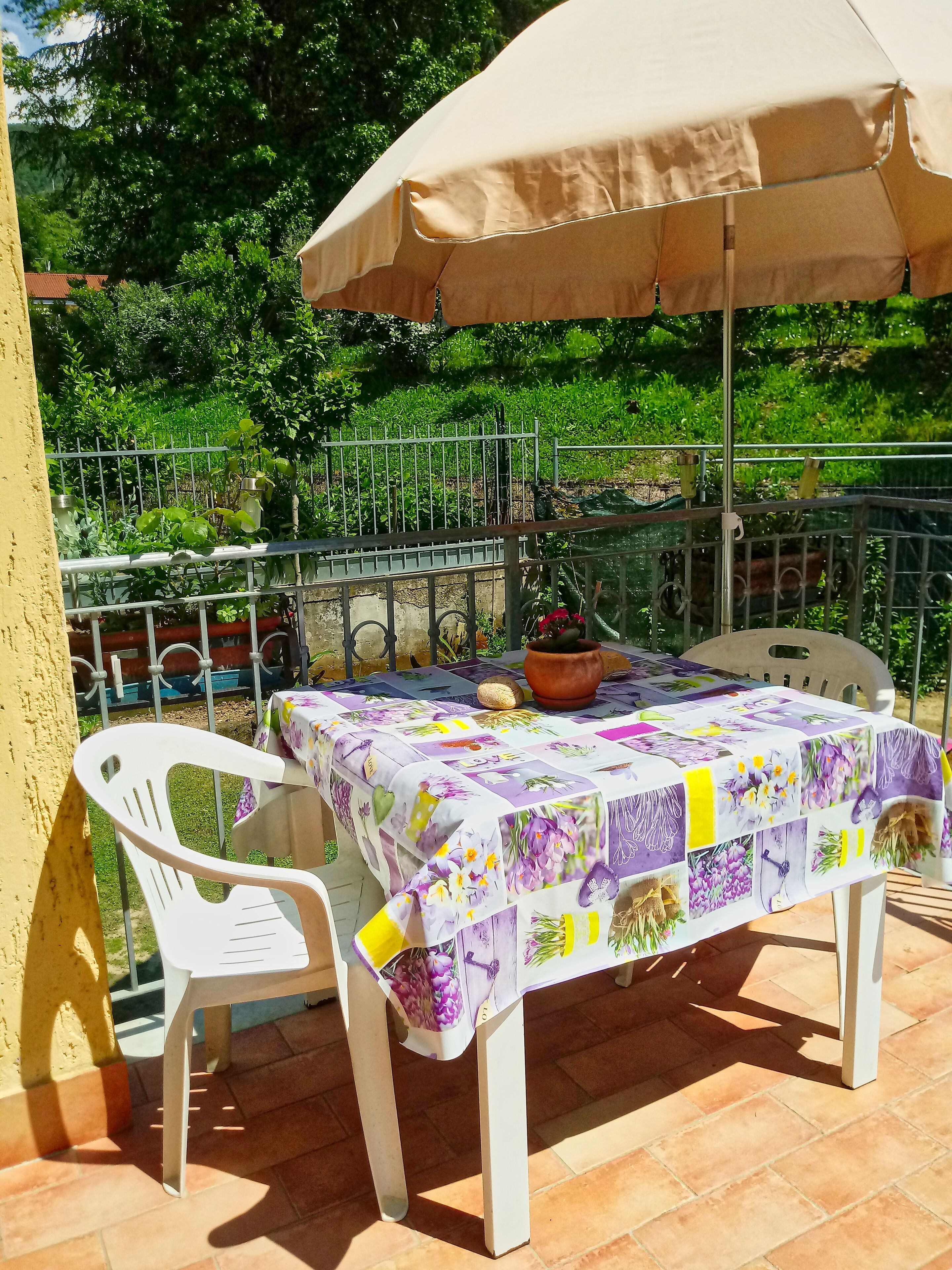 Terrassa a l'aire lliure amb una taula coberta amb un mantell de flors, una cadira de plàstic blanca i un para-sol beix que ofereix ombra.