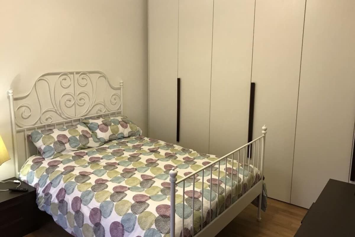 A bedroom with a metal bed frame, a patterned bedspread, a nightstand with a lamp, and a wardrobe with vertical handles.