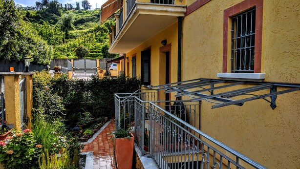 Ein gelbes Gebäude mit einem Balkon, einem Garten mit verschiedenen Pflanzen und Blick auf eine Hügelkuppe.
