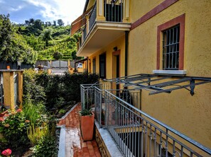 A yellow building with a balcony, a garden with various plants, and a view of a hillside. - Sally's B&B (Carrara)