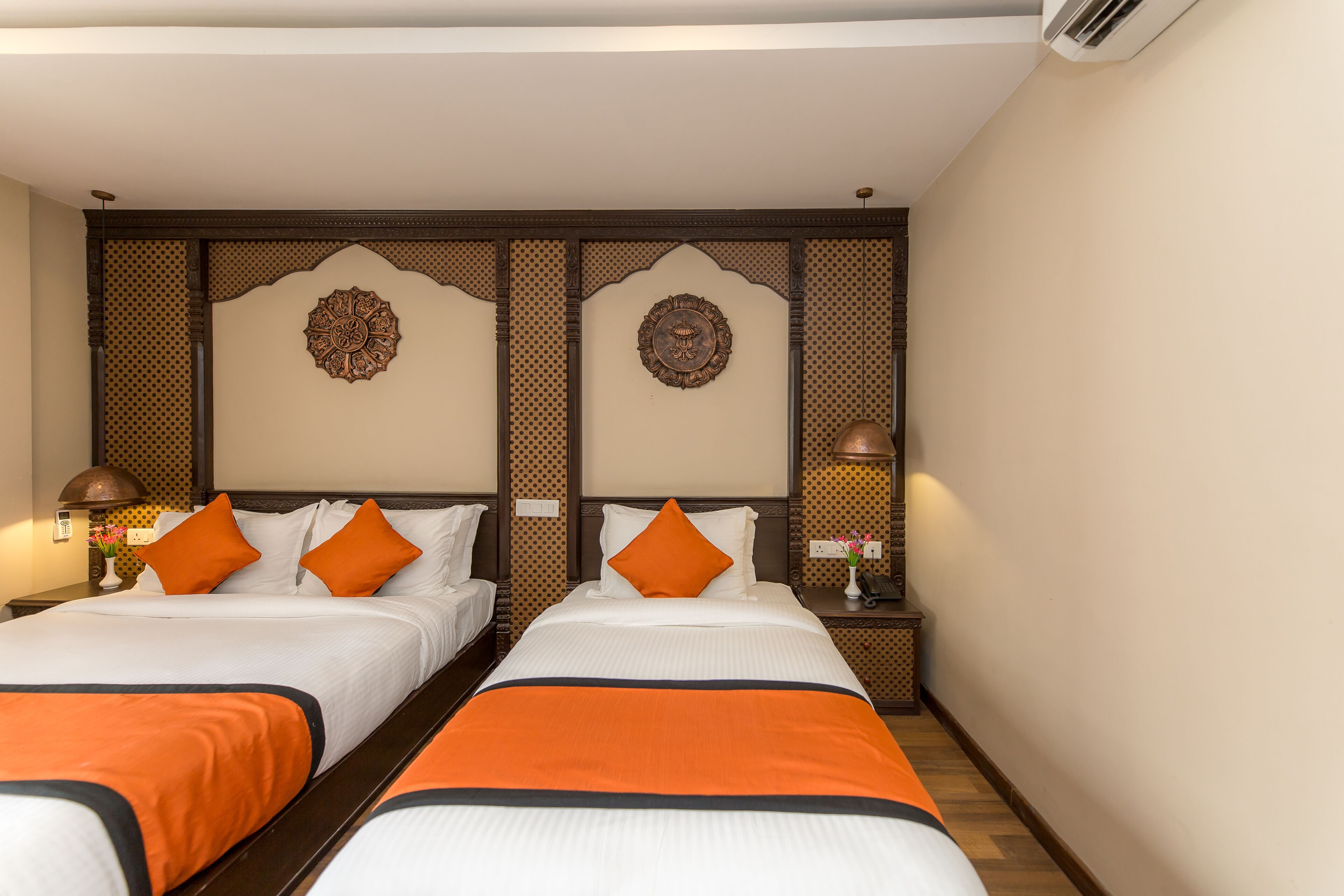 Hotelkamer met twee bedden, elk met witte beddengoed en oranje accenten. Er zijn decoratieve wandpanelen en een klein tafeltje met een lamp.