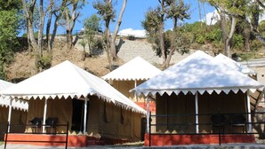 Une rangée de tentes blanches, dont les bases sont rouges, se dressent sur un fond de verdure composé d’arbres et d’une colline.