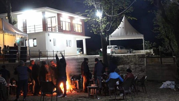 Une réunion en plein air autour d’un feu, avec des personnes assises à des tables, et un bâtiment en arrière-plan.