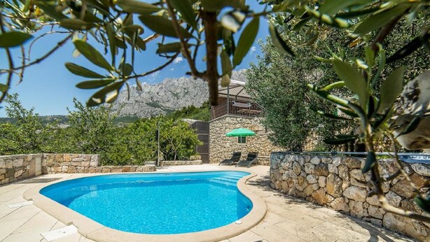 A swimming pool with a stone wall and lounge chairs, surrounded by greenery and a mountainous backdrop. - Villa Belvedere (Makarska)