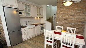 A compact kitchen with white cabinets, a stainless steel refrigerator, a microwave, and a stove. There is a dining table with white chairs and a red cushion. The floor is wooden, and there is a brick wall in the background. - Villa Belvedere (Makarska)