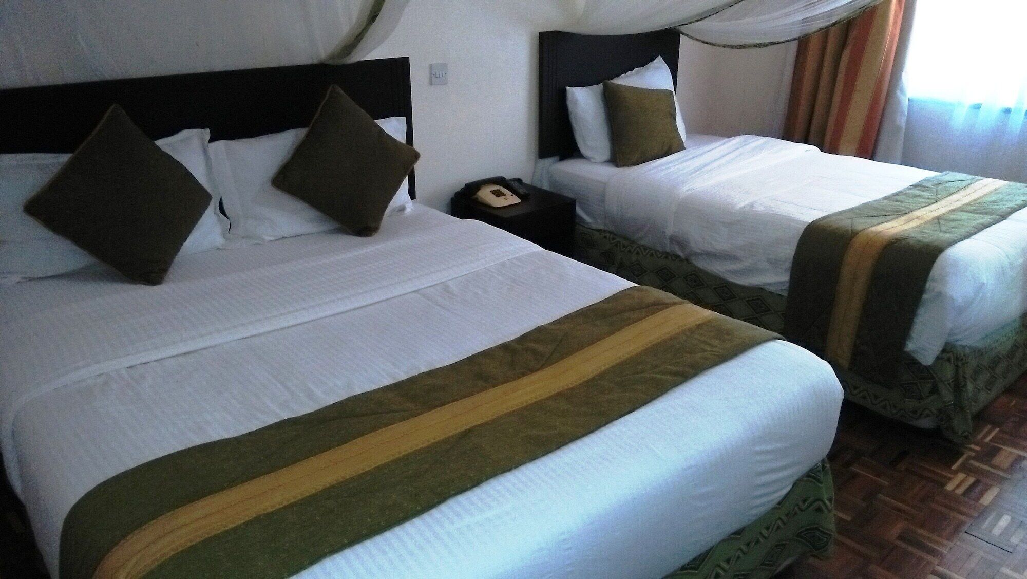 Two adjacent beds with white linens and green and brown striped bedspreads, a bedside telephone, and a curtain.