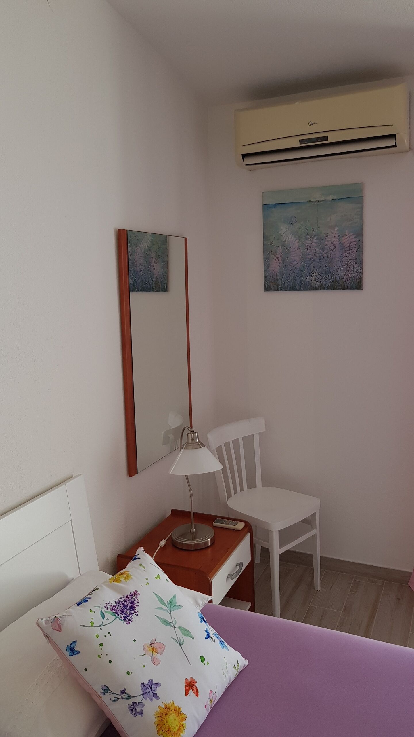 A bedroom with a bed, a desk with a lamp, a chair, a mirror, and two paintings on the wall.
