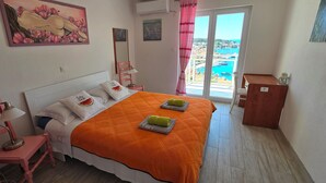 Un dormitorio con cama, una silla, un escritorio, una lámpara, un cuadro, un espejo y vista al mar a través de una ventana.