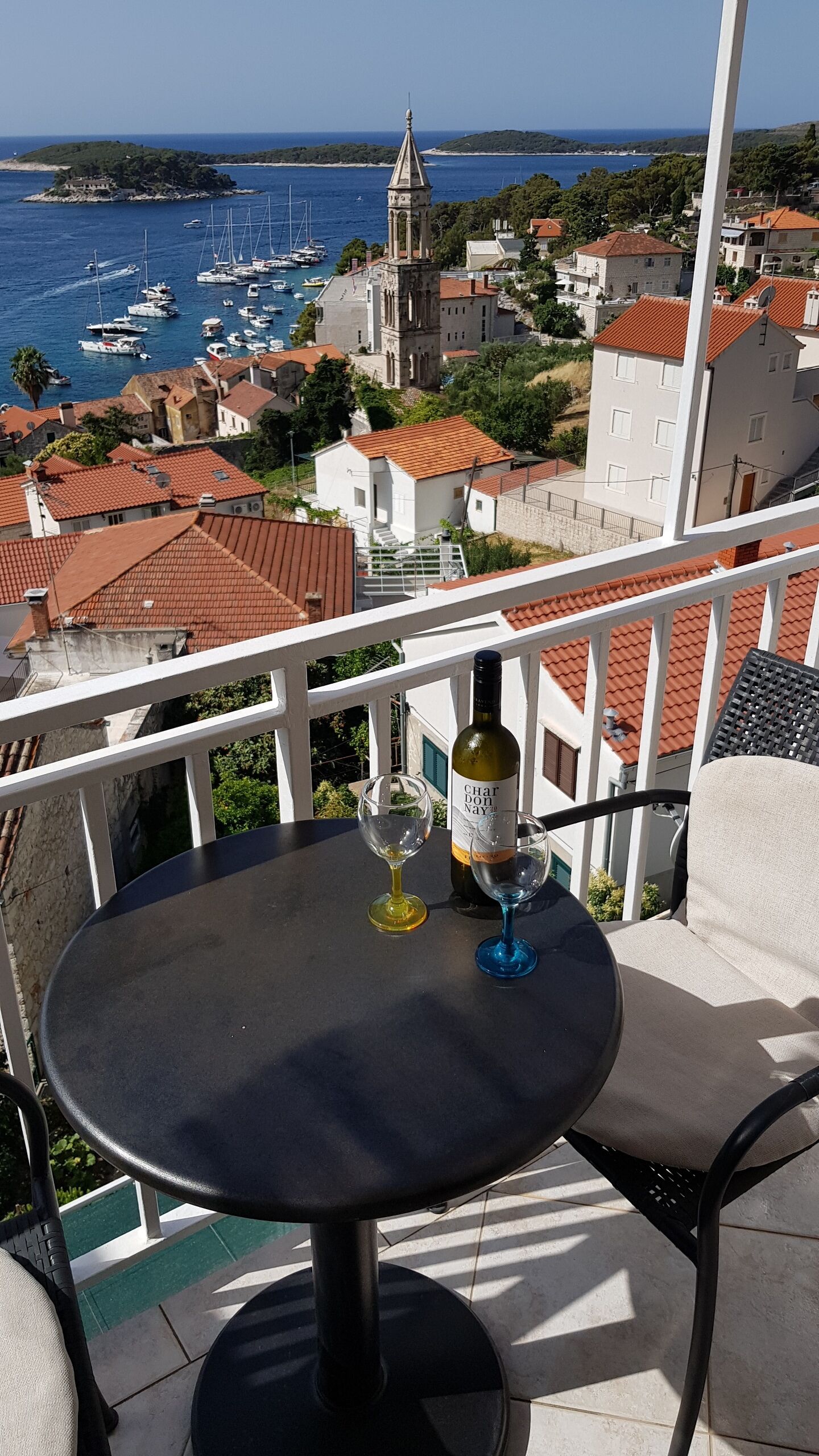 Un balcon avec une table, deux verres à vin et une bouteille de vin. La vue comprend un port de plaisance avec des bateaux et une église.