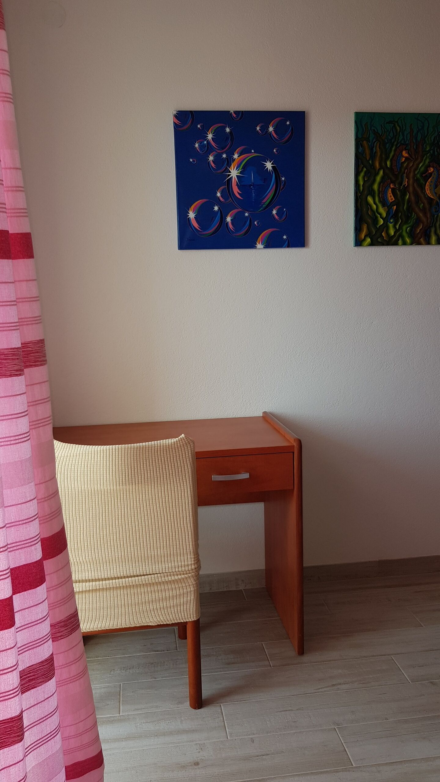 A room with a wooden desk, a wicker chair, and two abstract paintings on the wall.