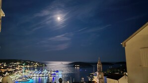 Vista nocturna de un pueblo costero con puerto deportivo, barcos y una torre de iglesia destacada.