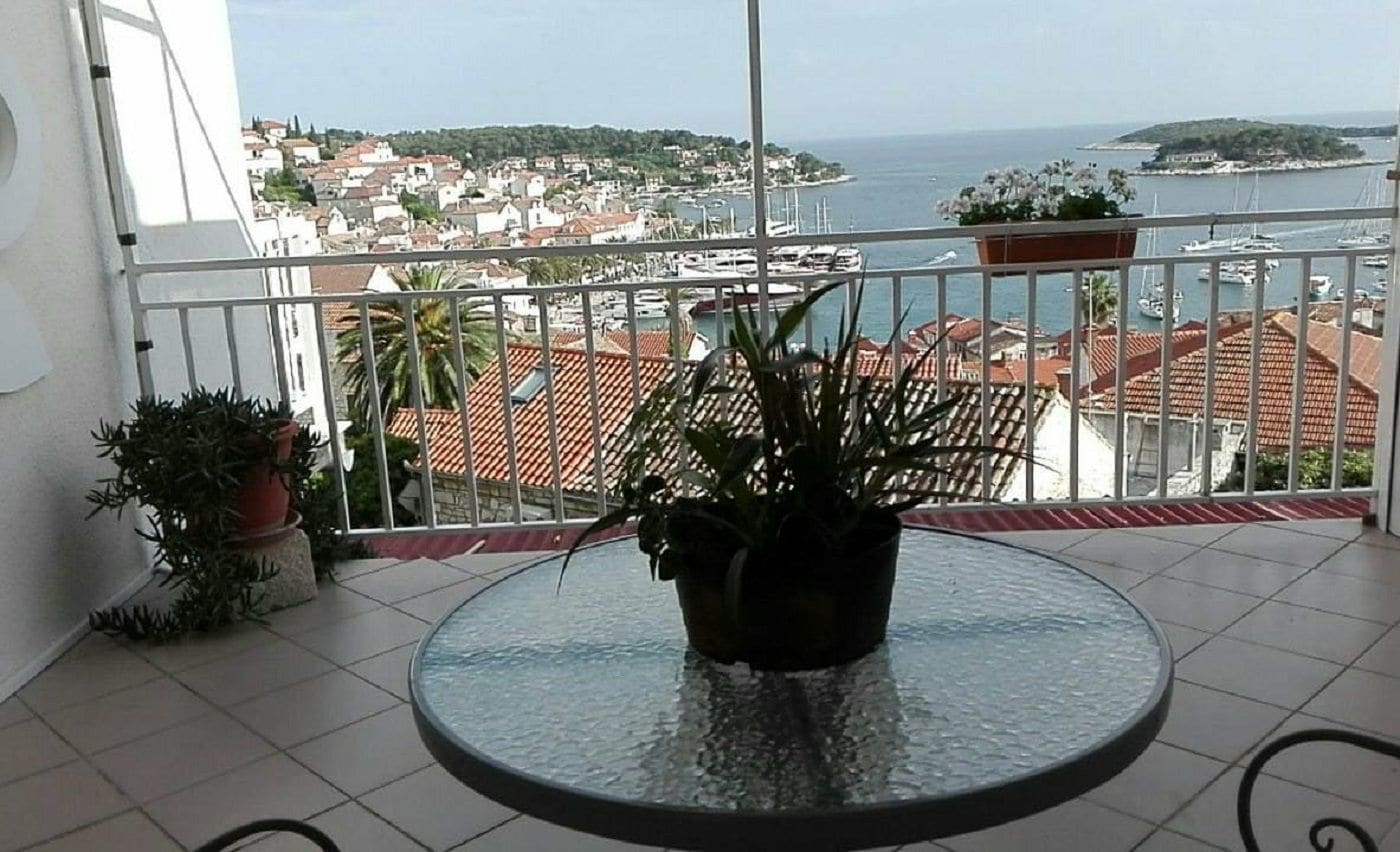 A balcony with a round glass table, potted plants, and a view of a coastal town and marina.