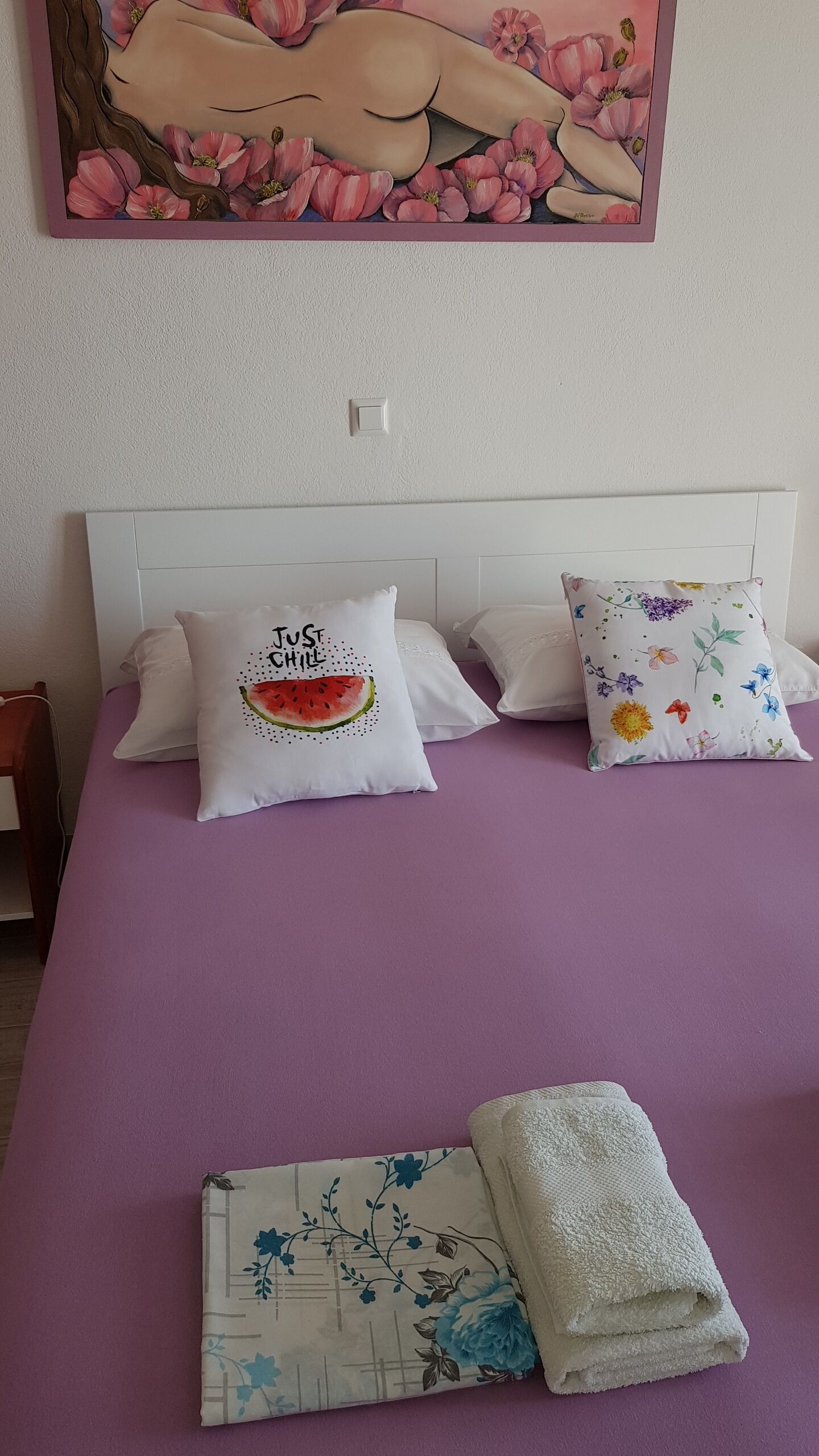 En seng med lilla dyne, to hvide puder med blomstermønstre, og et billede af en kvinde med blomster på væggen over sengen.