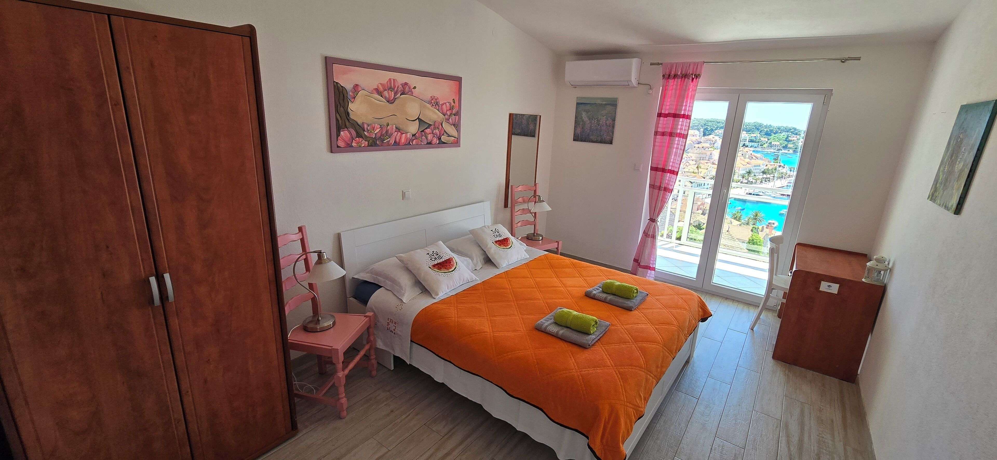 A bedroom with a bed, a nightstand, a wooden wardrobe, a window with a view, and a painting on the wall.