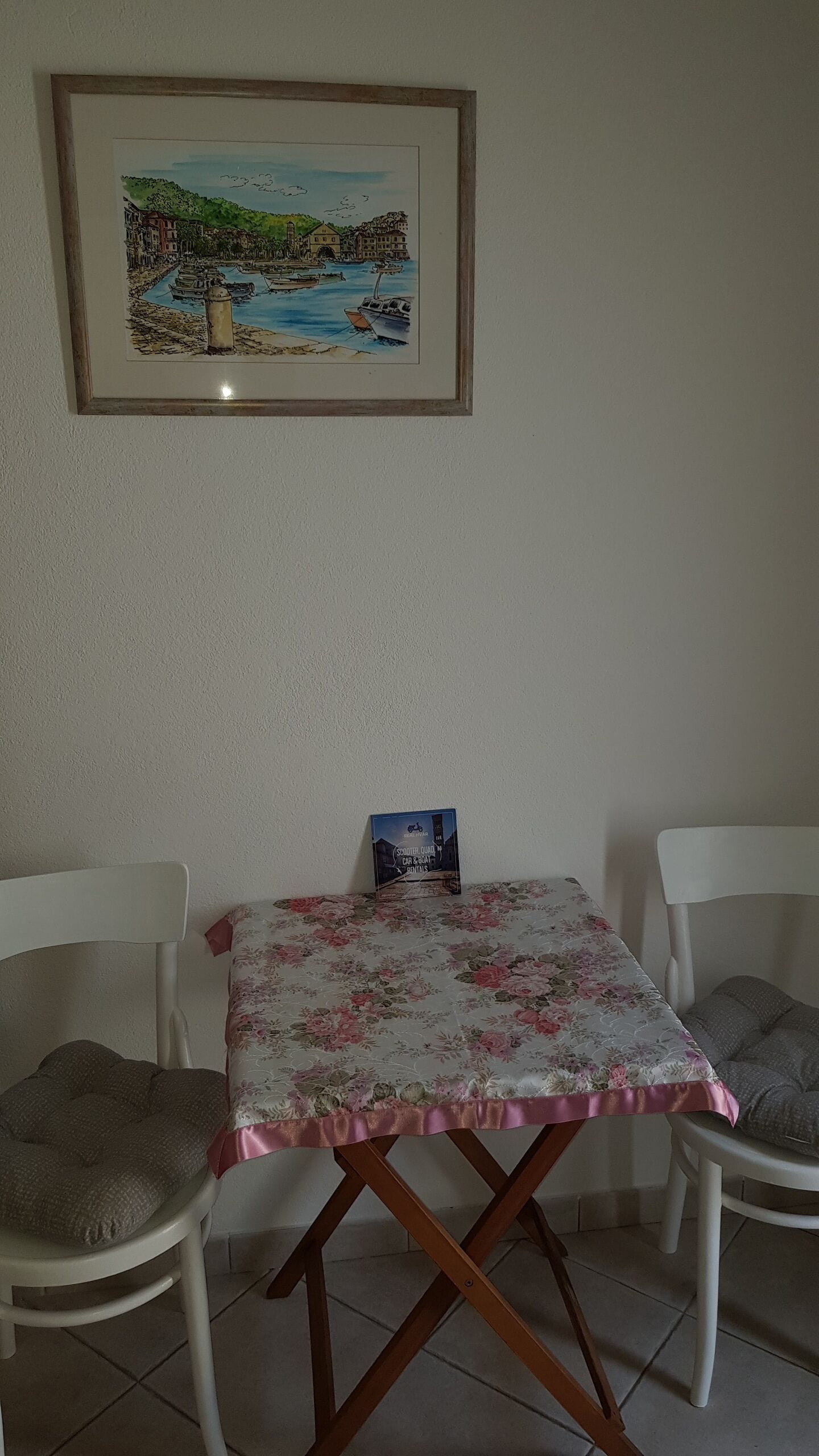 A dining area with a table covered by a floral tablecloth, two white chairs, and a framed painting above.