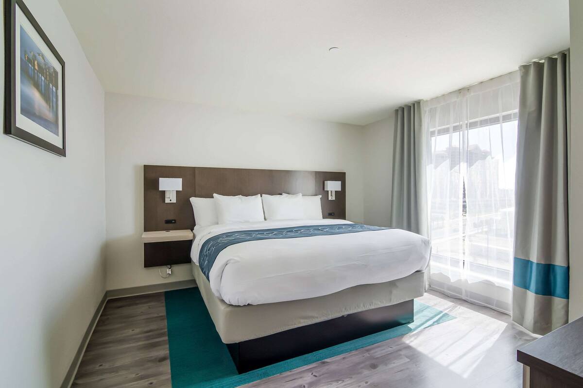 A hotel room with a large bed, a nightstand, a lamp, and a view of the city through sheer curtains.