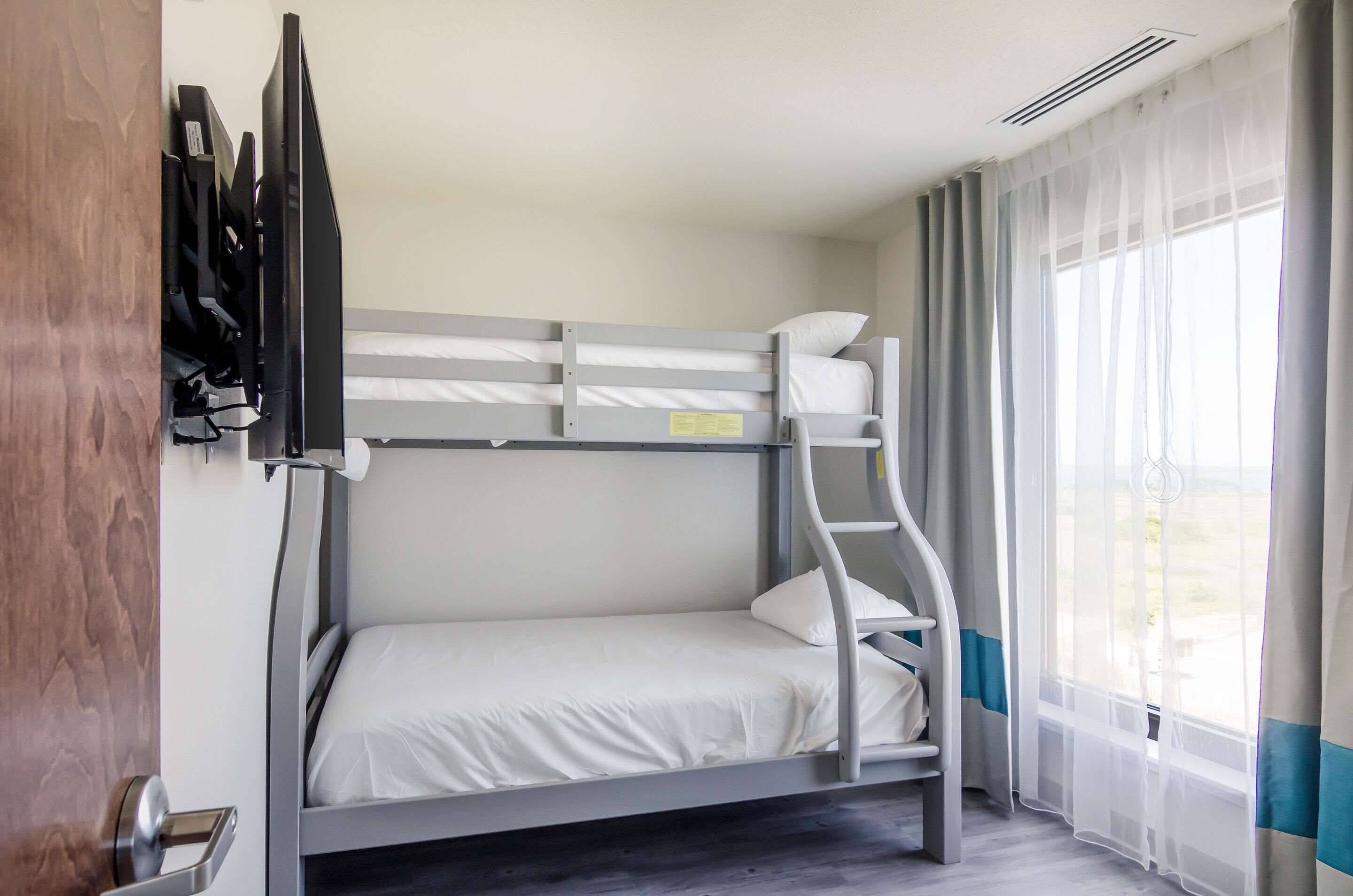 A bunk bed room with a TV mounted on the wall, a window with sheer curtains, and a wooden door.